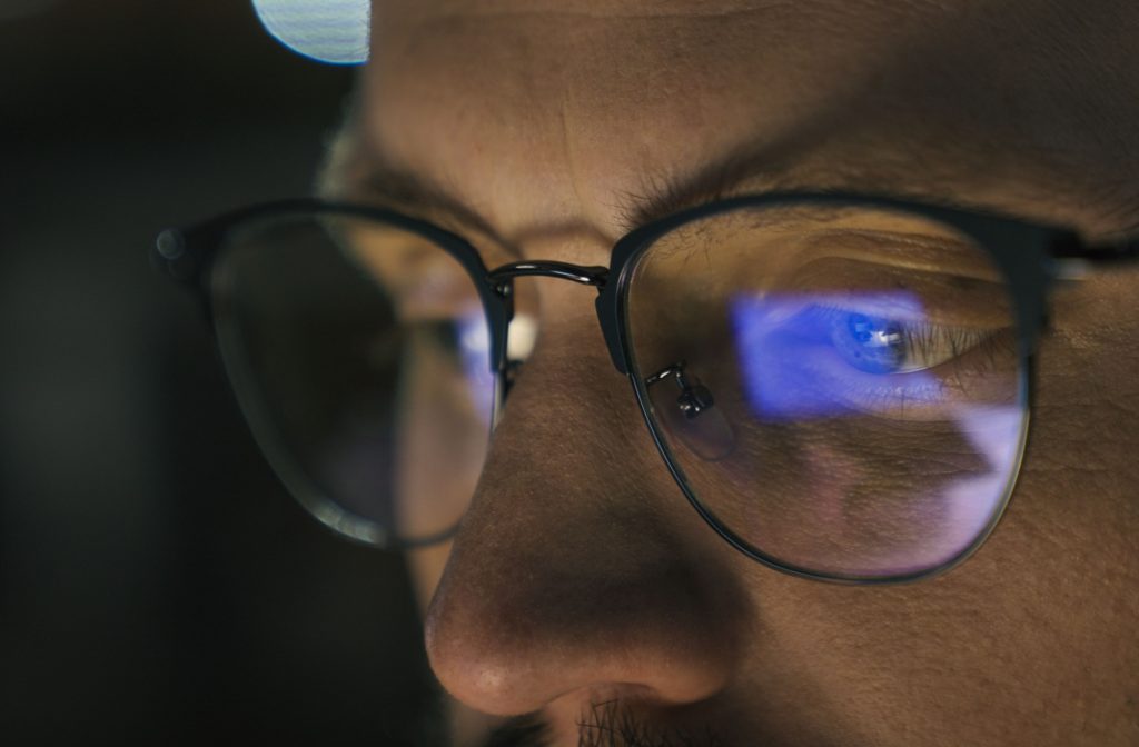 A close-up of a person's upper face as they work on a computer at night. The reflection of the screen is visible in the lenses of their glasses.