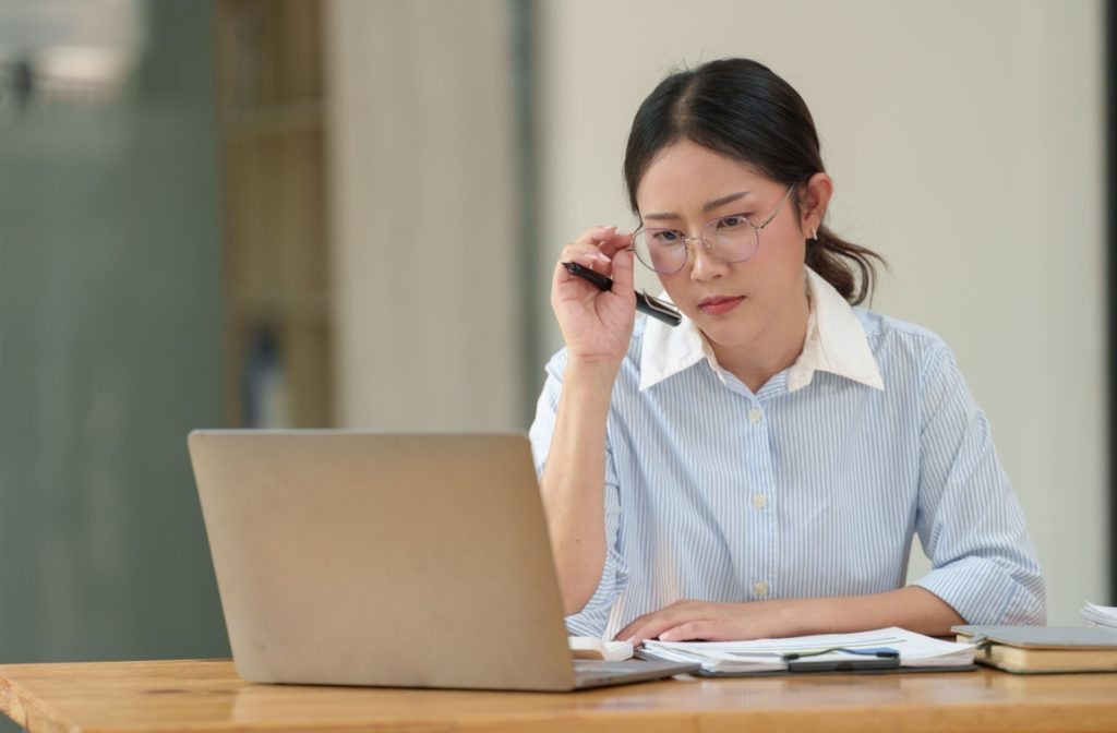 An office worker sits at a table, adjusting their glasses while they work on their laptop. The laptop is approximately arm's length away from them.