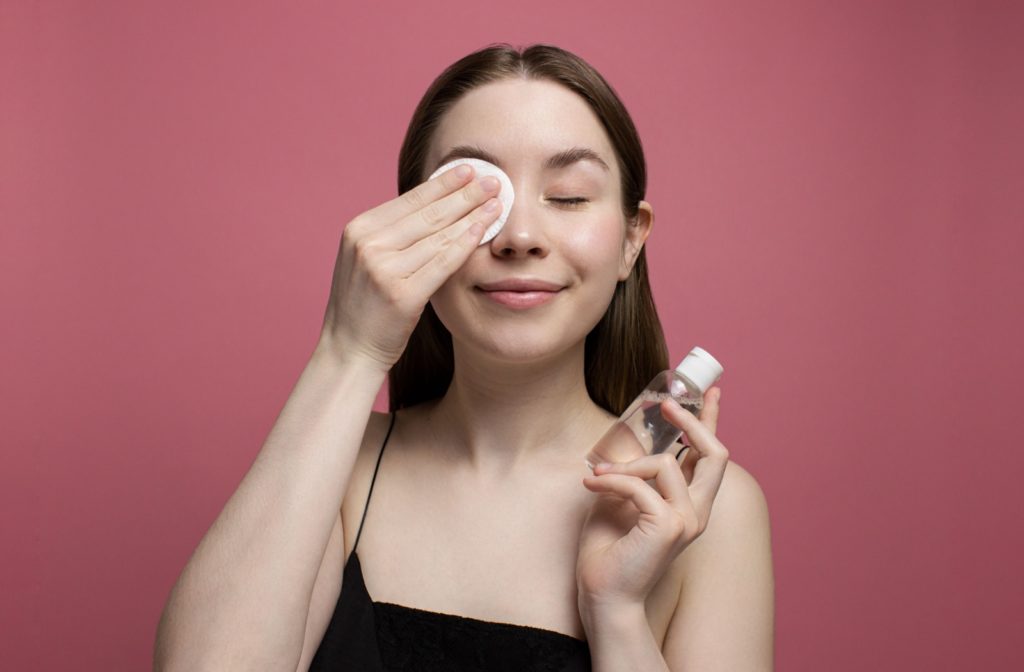 A young adult against a pink background smiles as they clean their eyelids with unscented soap