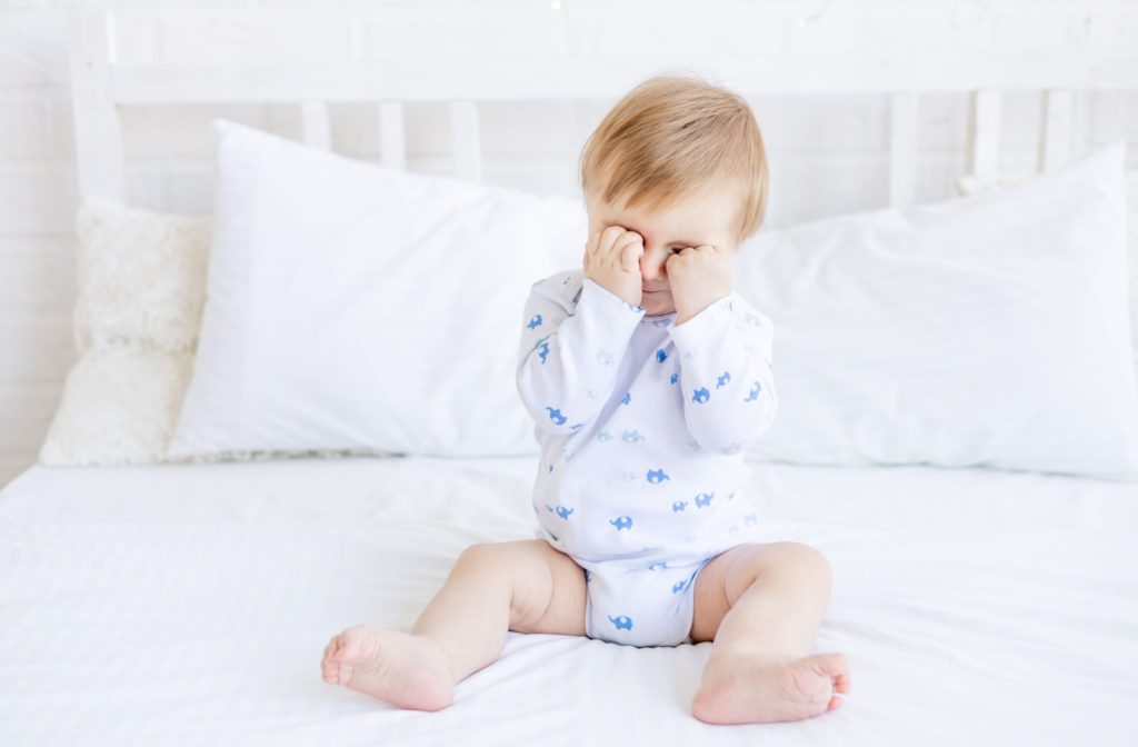 A baby sitting on a bed rubbing their eyes with both hands.